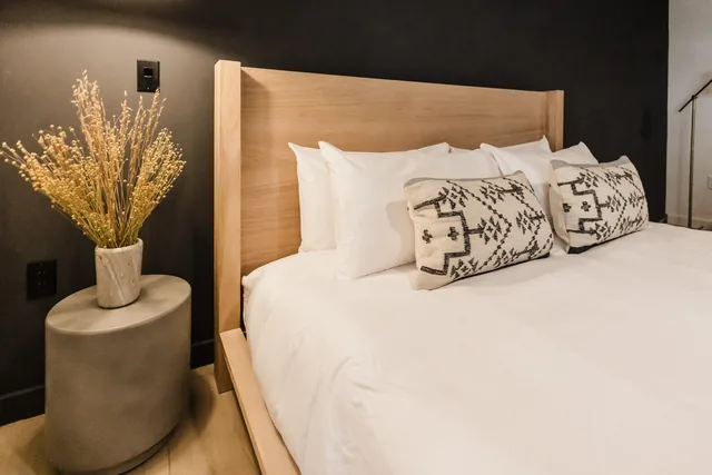 Natural oak headboard with white linens, woven geometric lumbar pillows, and dried botanicals on concrete nightstand