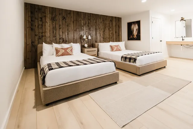 Spacious double-queen room with wood accent wall, white linens, plaid throws, and open vanity alcove beyond