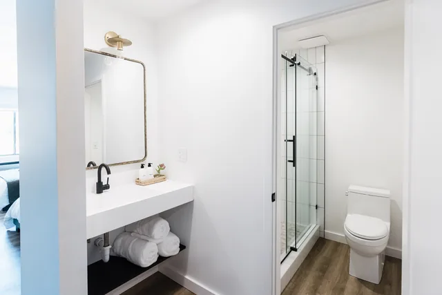 Spotless white bathroom with brass sconce, black fixtures, glass-enclosed shower, and rolled towels