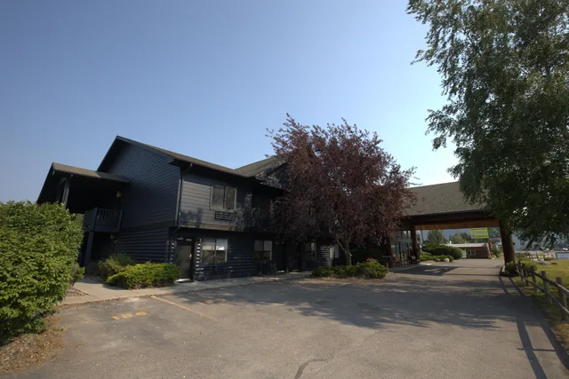 Dark-painted hotel exterior with covered drive and ornamental trees under a clear Montana sky