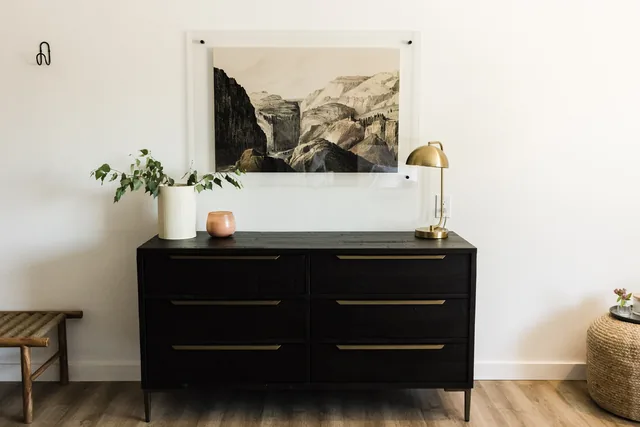 Black wood dresser with brass lamp, white ceramic vase with greenery, and a Glacier canyon art print in an acrylic frame