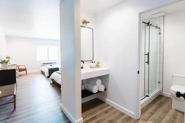 Open-plan double queen room seen through the bathroom partition, showing floating vanity with rolled towels and glass-door shower