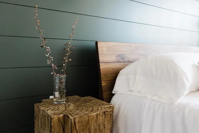 Live-edge tree-stump nightstand with wildflower sprig in glass vase beside crisp white king pillows