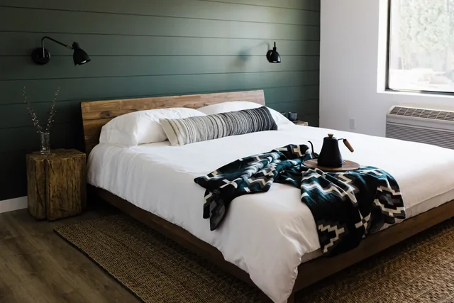 King bed with Pendleton-style throw, pour-over kettle on wood tray, and warm sconce lighting against green shiplap