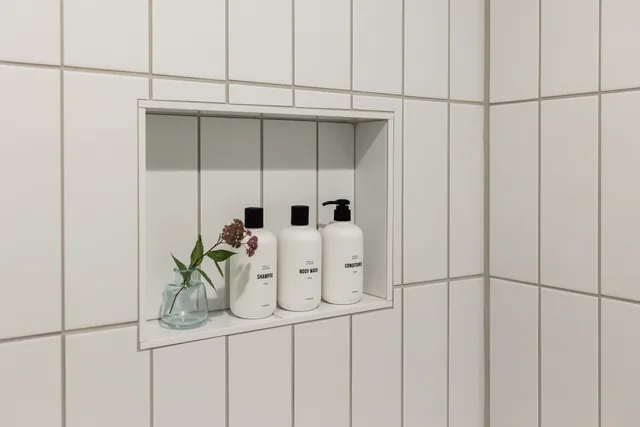 Built-in shower niche with branded shampoo, body wash, and conditioner bottles beside a small wildflower vase on white subway tile
