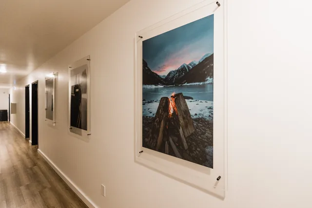 Hotel corridor lined with acrylic-mounted local photography, the foreground print showing a campfire on a glacial lakeshore at dusk