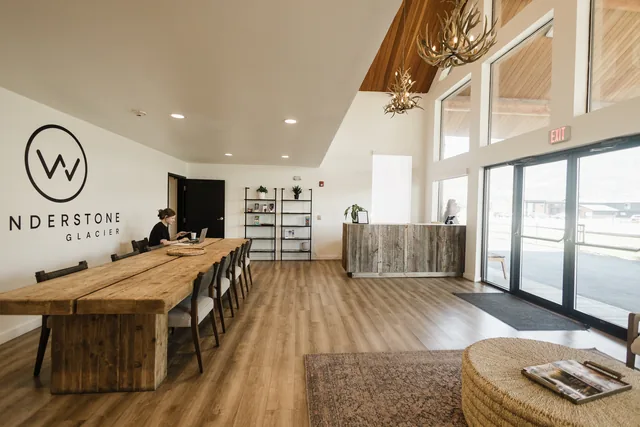 Wonderstone Glacier lobby — vaulted wood ceiling, antler chandelier, reclaimed-wood front desk, and communal farm table bathed in mountain light