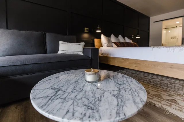 Marble coffee table with candle in foreground, sofa and king bed with lit sconces softly focused behind